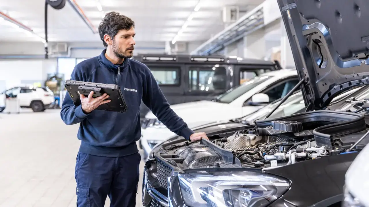 Meccanico con tablet analizza la differenza tra tagliando e revisione auto, con cofano aperto.