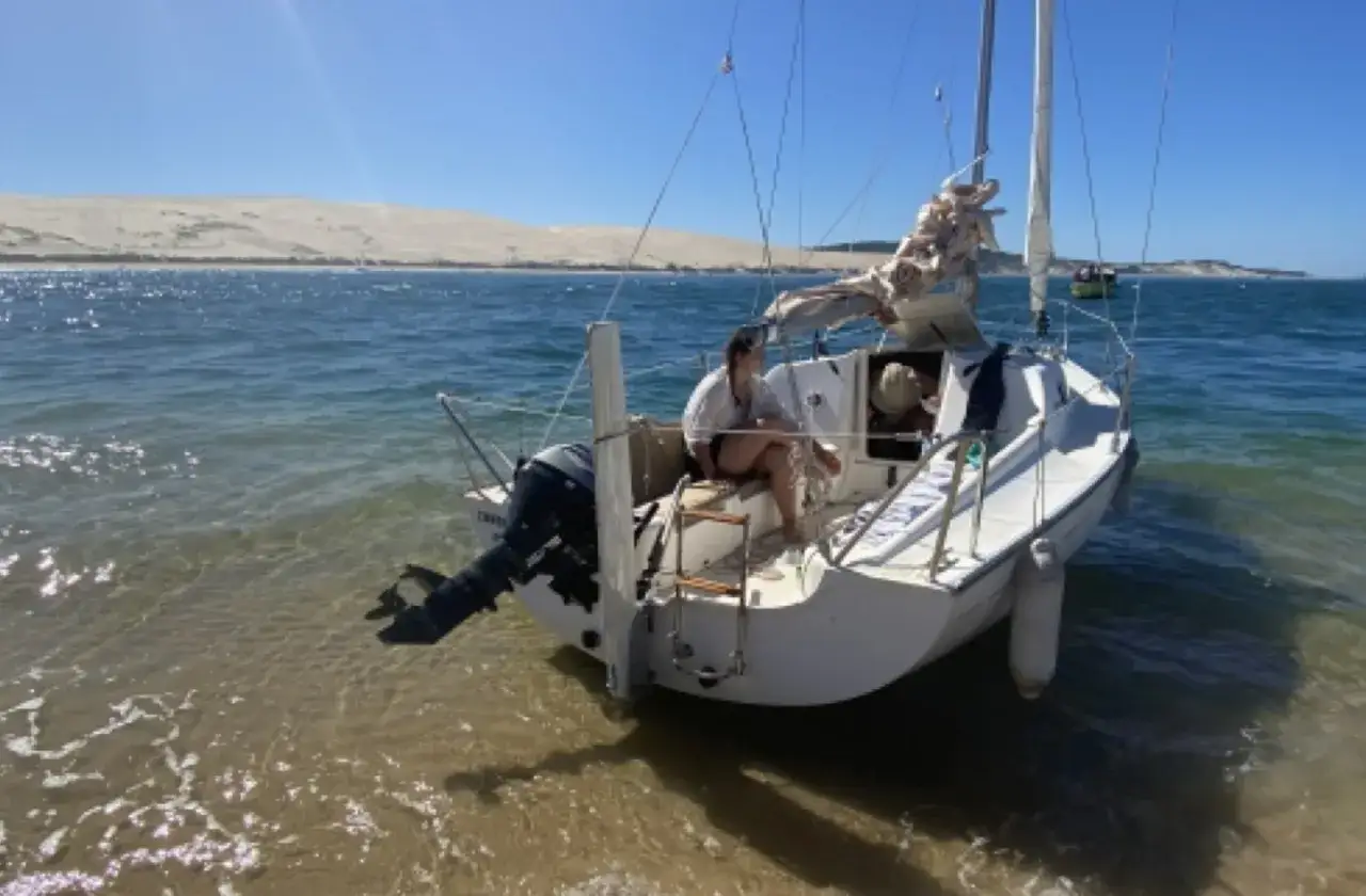 Voilier Sun 2000 échoué sur une plage de sable, avec deux personnes à bord. Le soleil brille sur l'eau bleue.