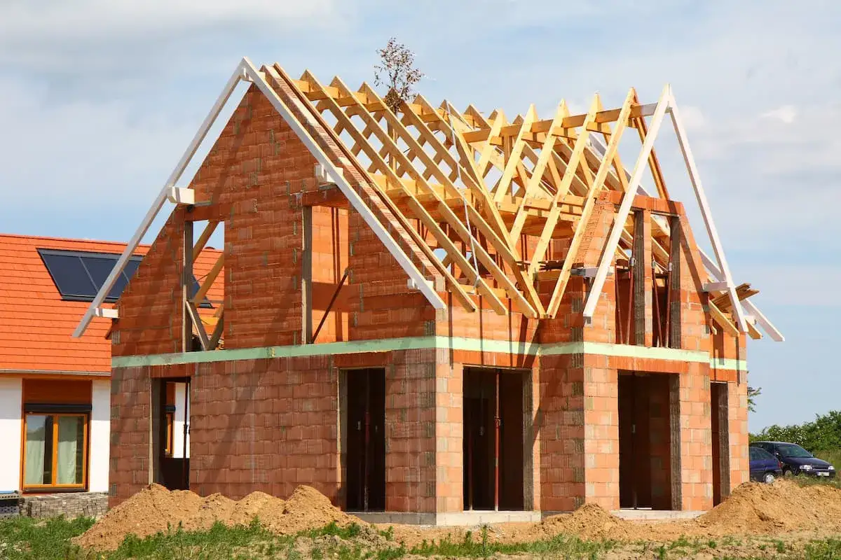 Rohbau firma baut ein Haus aus Ziegeln mit sichtbarem Dachstuhl. Daneben ein fertiges Haus mit roten Ziegeln.