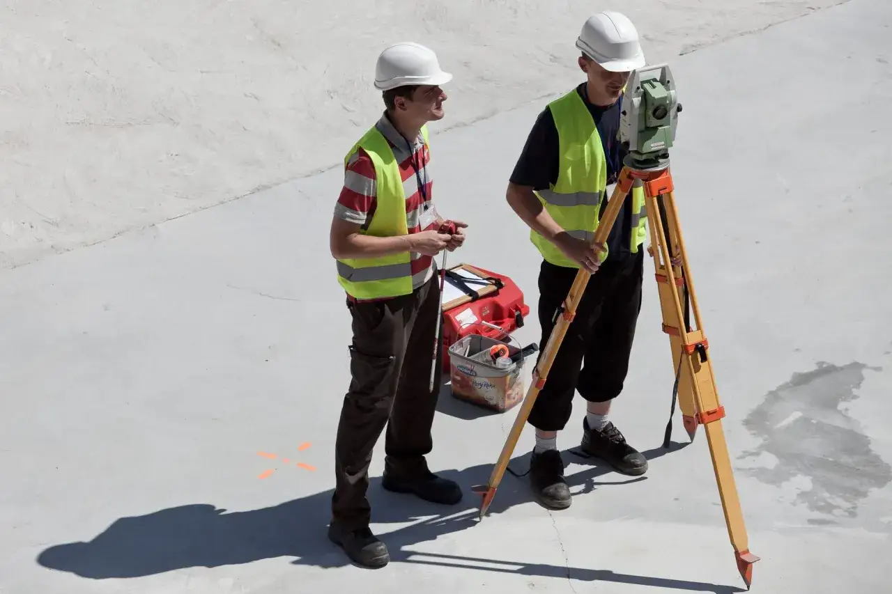 Dwóch geodetów w kamizelkach odblaskowych i kaskach pracuje z niwelatorem. Zastanawiasz się, ile zarabia geodeta?