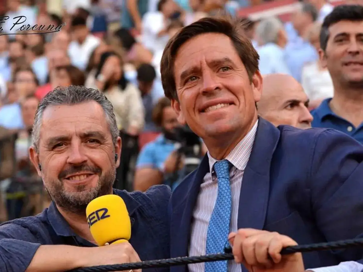 Eduardo D&aacute;vila Miura, sonriente, con traje azul, junto a un periodista con micr&oacute;fono amarillo en una plaza de toros.