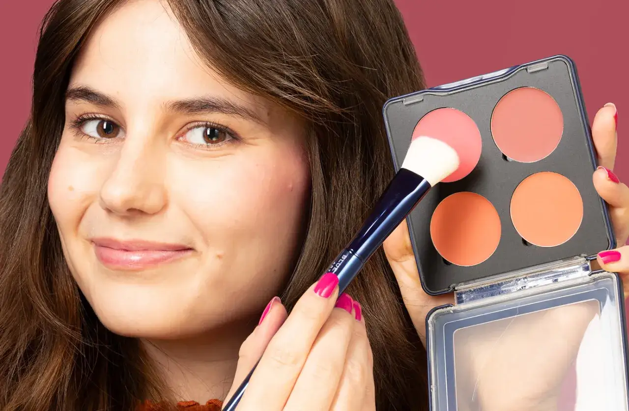 Una mujer sonríe mientras aprende como aplicar colorete con brocha, usando una paleta de rubores en tonos rosados y naranjas.