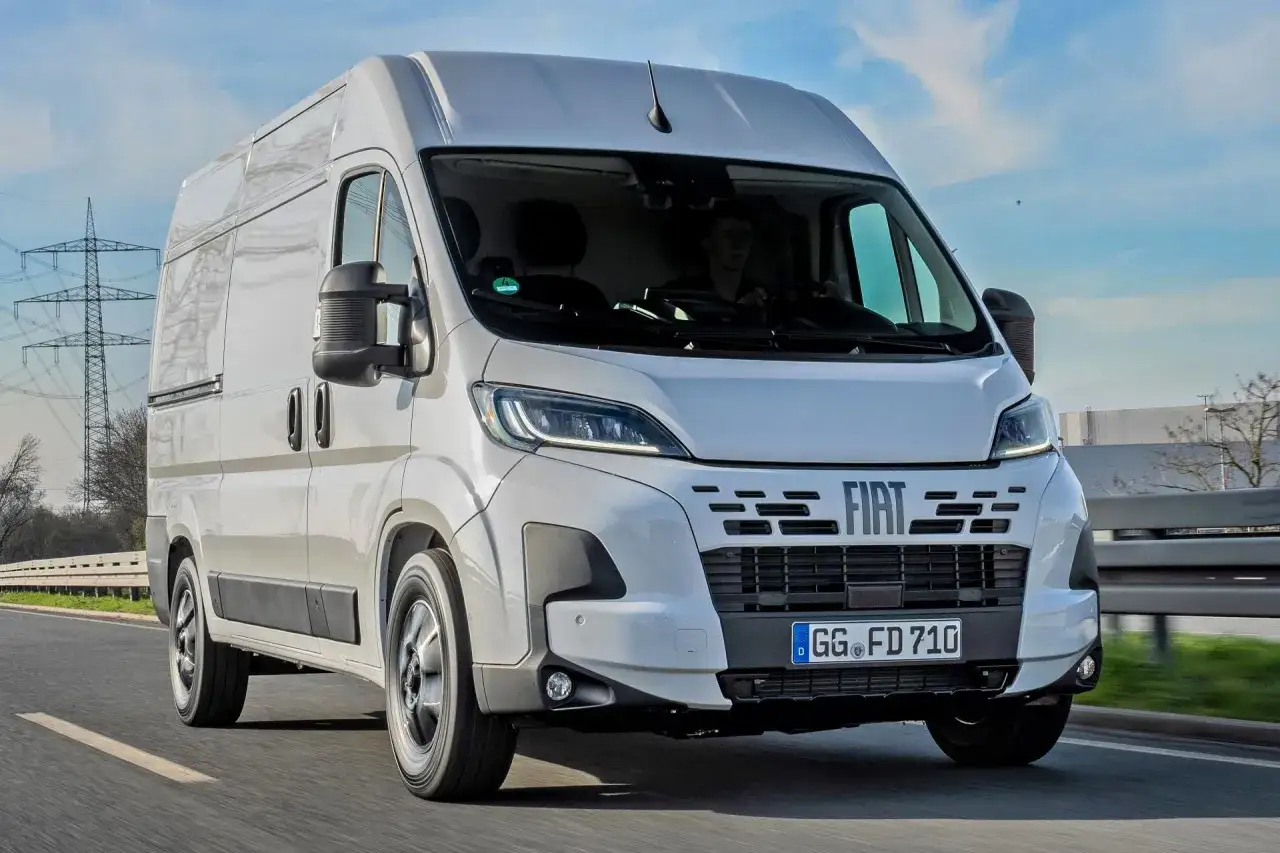 Biały Fiat Ducato Maxi w ruchu drogowym, z kierowcą za kierownicą, na tle słonecznego nieba i linii energetycznych.