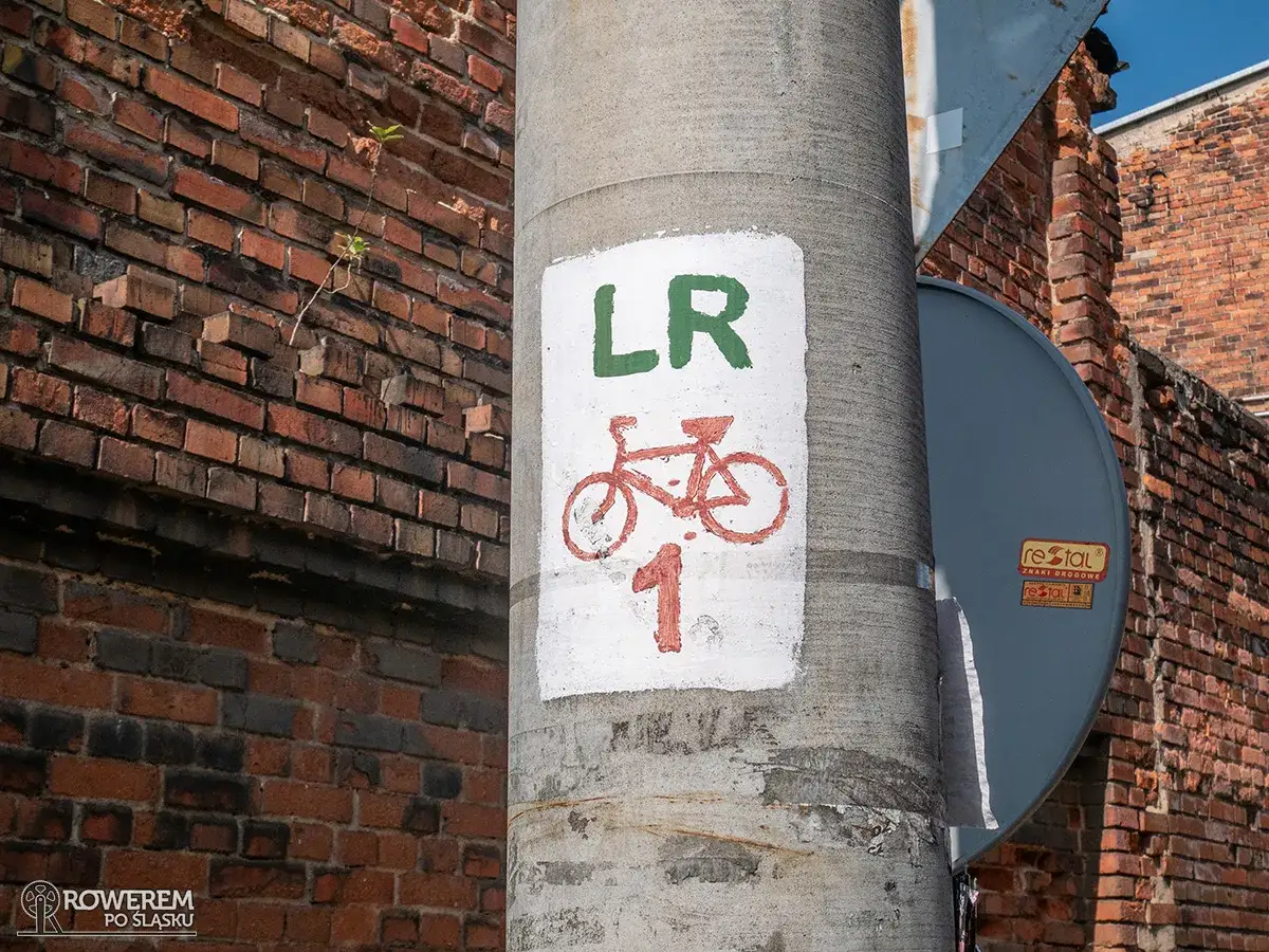 Znak na słupie informuje o trasie rowerowej LR 1. Czerwony rower i numer wskazują drogę dla miłośnik&oacute;w dw&oacute;ch k&oacute;łek.