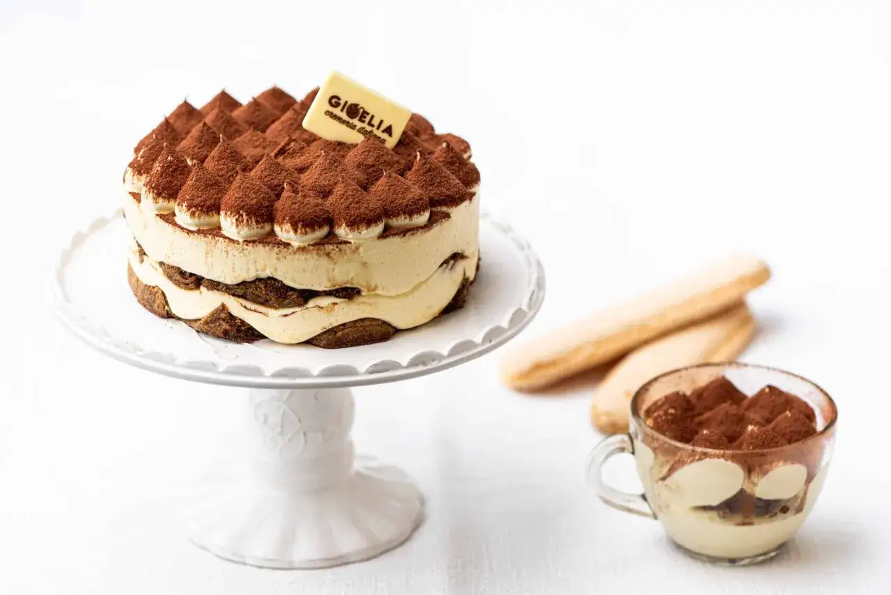 Pastel de tiramisú con crema y cacao, acompañado de bizcochos de soletilla y una copa de postre. Los tiramisú ingredientes perfectos para un deleite.