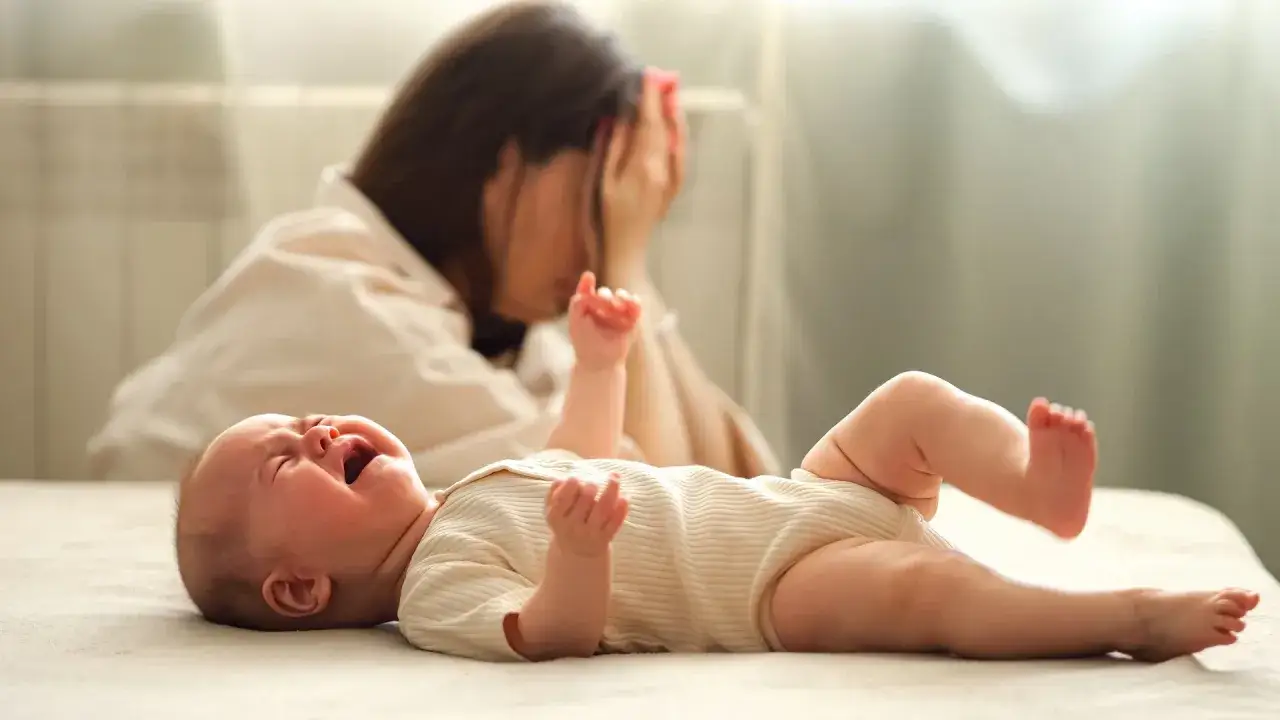 Verzweifelte Mutter h&auml;lt sich den Kopf, w&auml;hrend ihr schreiendes Baby mit scheinbarer Hochbegabung nach Aufmerksamkeit verlangt.