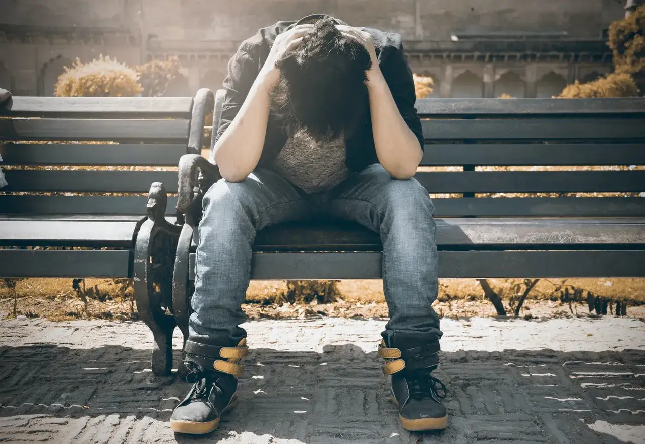 Mężczyzna siedzi na ławce, trzymając się za głowę. Jego postawa sugeruje cierpienie, być może związane z psychozą amfetaminową.