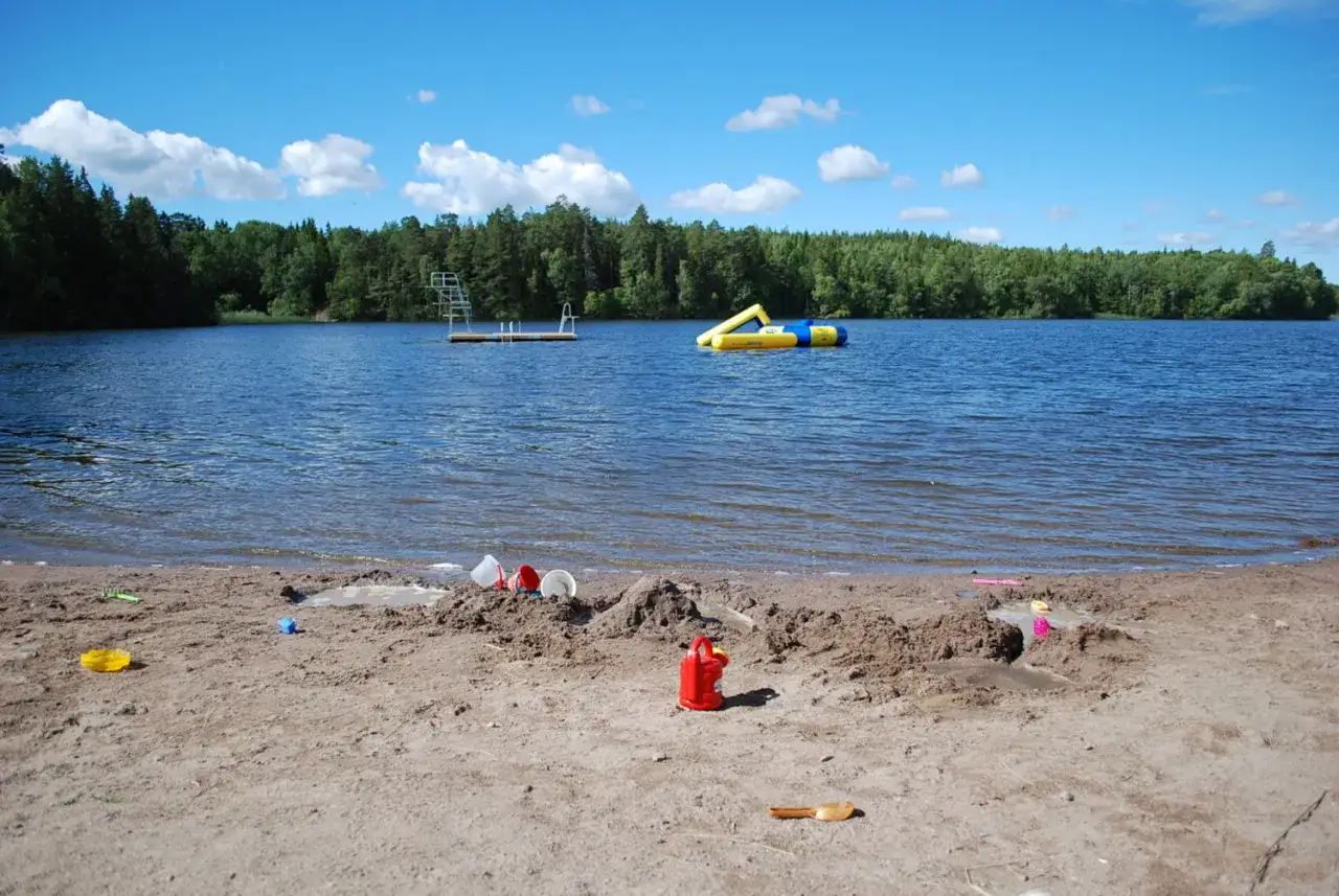 En solig dag vid en sjö med en badbrygga och en uppblåsbar vattenlek. Sandleksaker ligger utspridda på stranden, redo för lek. Kanske en dag för att bygga sandslott!