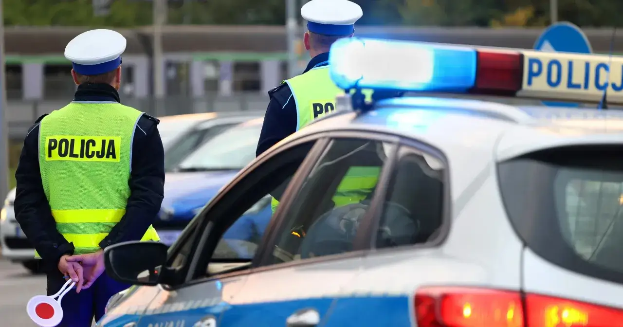 Policjanci w kamizelkach odblaskowych przy radiowozie z włączonymi sygnałami świetlnymi. Może to oznaczać kontrolę drogową lub zabranie prawa jazdy.