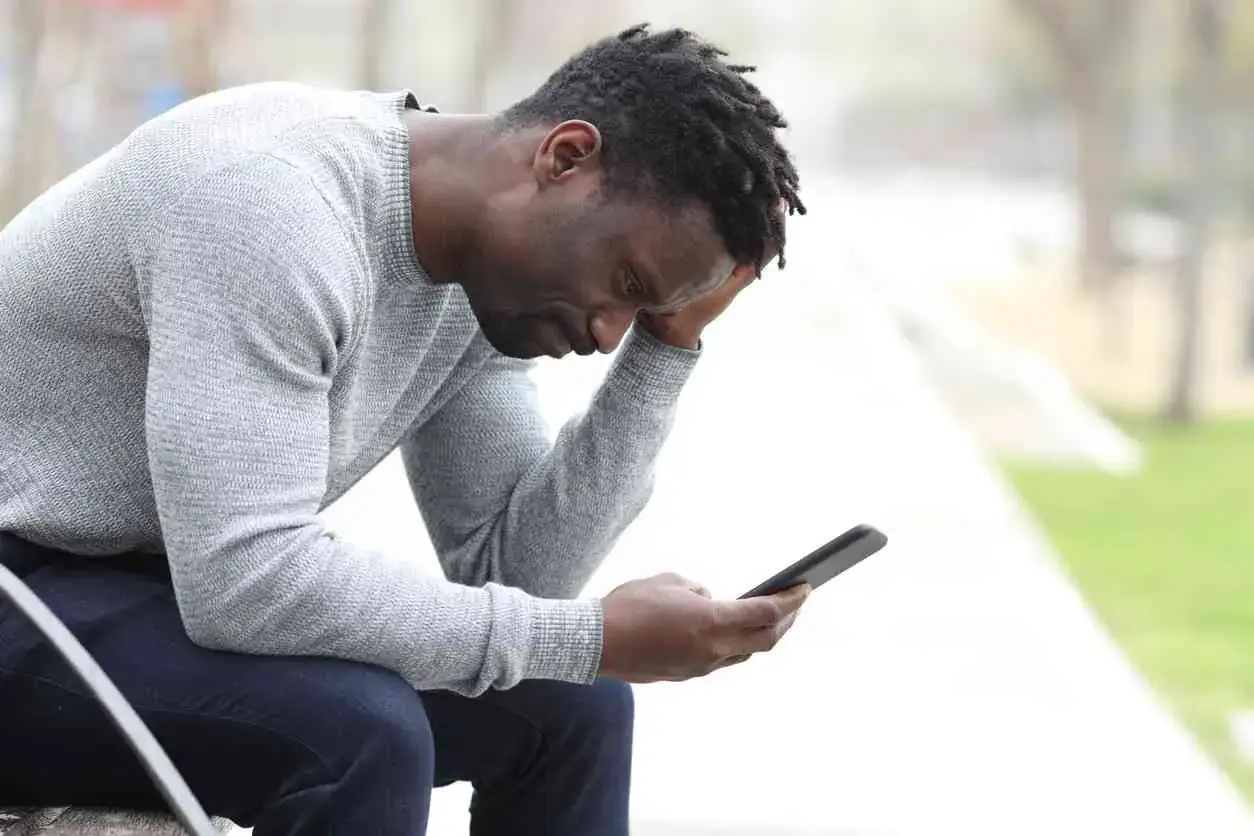 Uomo con dreadlocks seduto all'aperto, con la mano sulla fronte, che guarda il telefono con espressione angosciata. Sembra provare una forte paura di parlare al telefono.