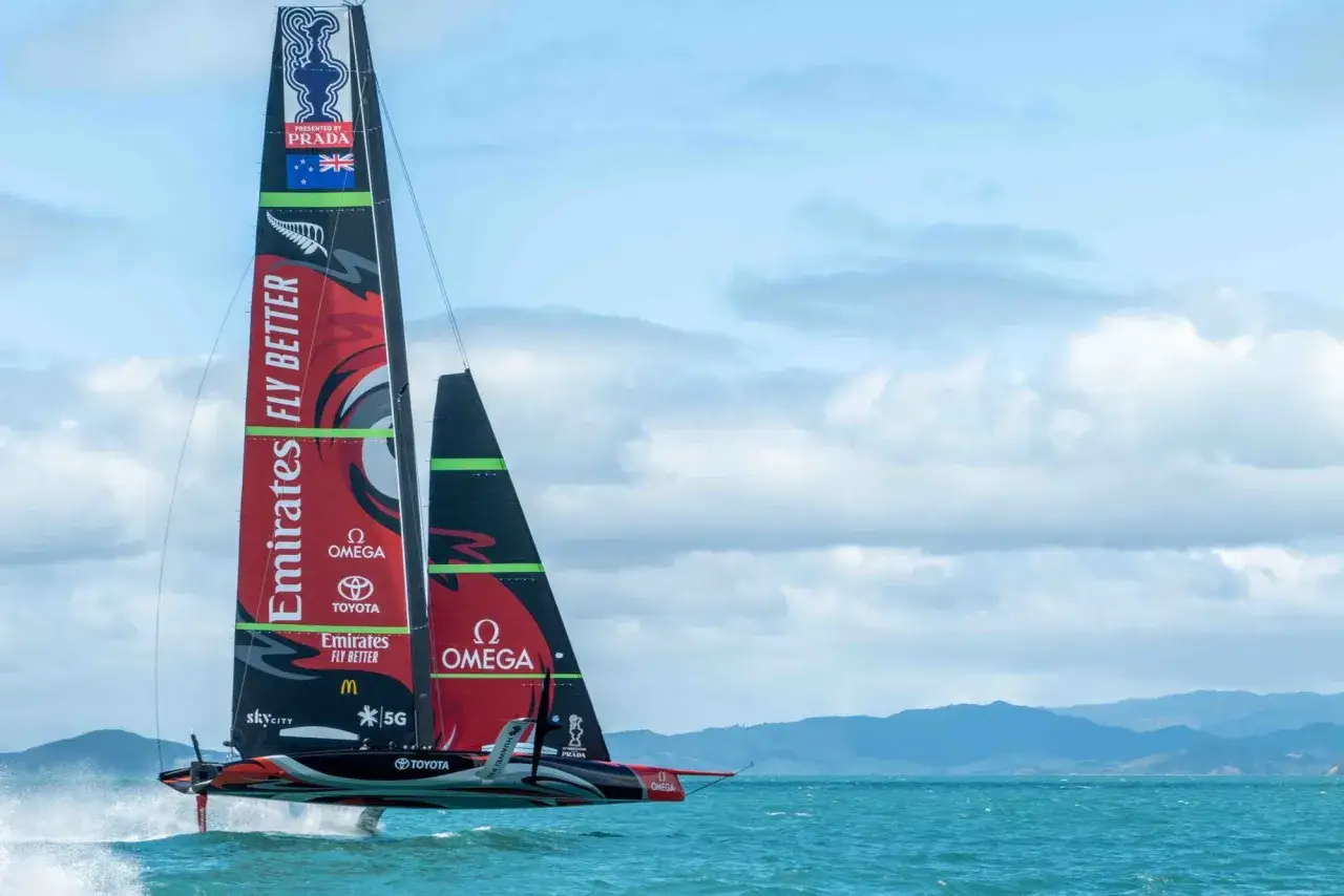 Barca a vela in foil che sfreccia sull'acqua, mostrando la sua incredibile velocit&agrave; massima. Le vele sono decorate con sponsor come Emirates e Omega.