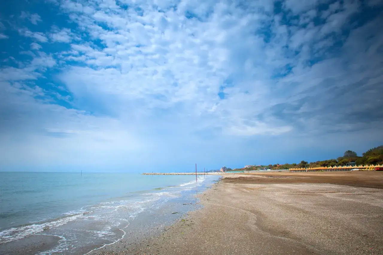 Ein weiter, ruhiger Strand am Lido di Venezia unter einem dramatischen Wolkenhimmel. Die Wellen des Meeres umspülen den sandigen Strand.
