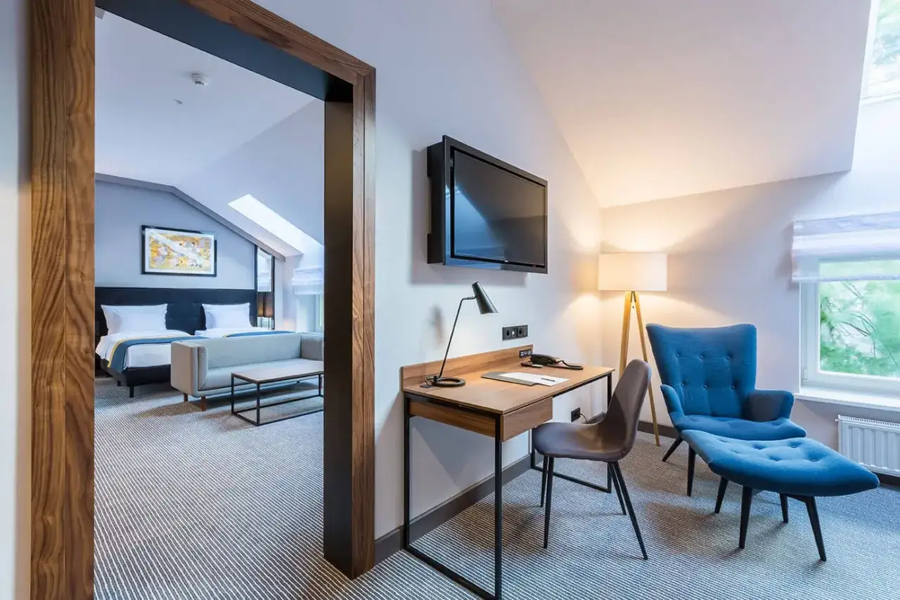 Pokój w hotelu 10 gwiazdkowym: łóżka, sofa, biurko z lampką i fotelem, telewizor.
