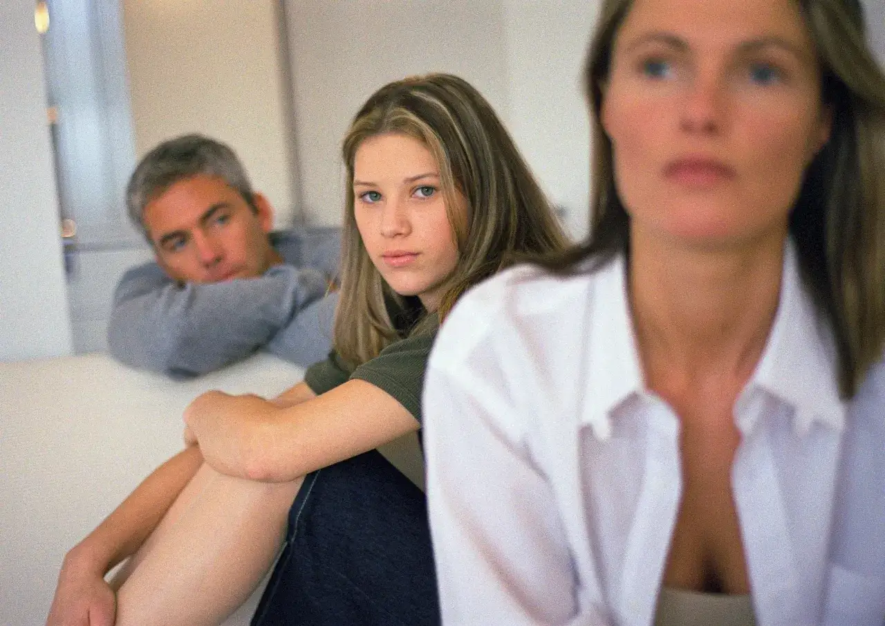 Ein Mann blickt nachdenklich, während ein Mädchen, etwa 12 Jahre alt, und eine Frau im Vordergrund sitzen.