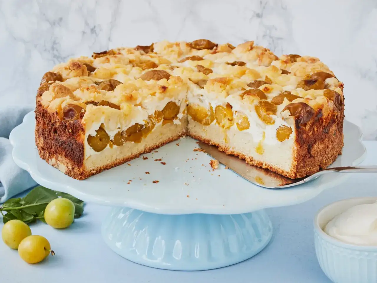 Saftiger Mirabellenkuchen mit Quark und Streuseln auf einem blauen Tortenständer. Frisch gebacken und bereit zum Genießen.