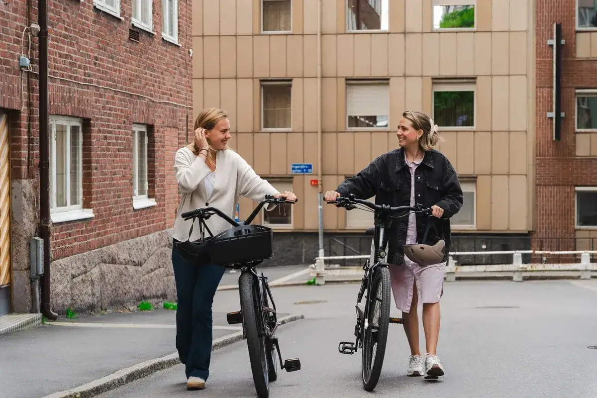 Två glada kvinnor går med sina elcyklar i en stadsmiljö. Kanske har de precis fått bidrag elcykel och njuter av sin nya frihet.