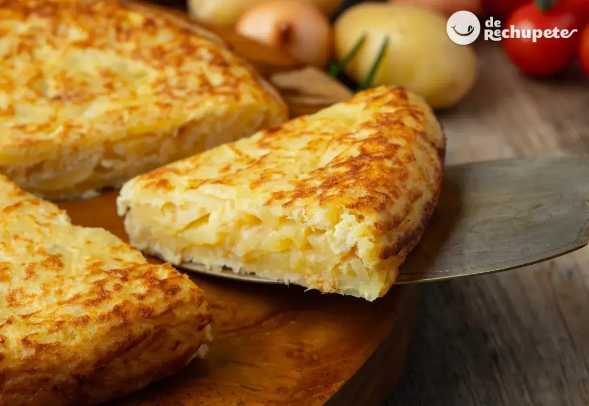 Una porci&oacute;n de tortilla de patatas dorada y jugosa se sirve sobre una pala. Patatas y cebollas en el fondo.