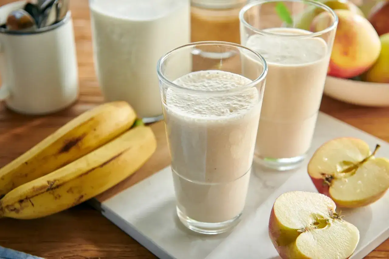 Dwa szklanki puszystego napoju, obok dojrzałe banany i jabłka. Idealny sok z banana na orzeźwienie.