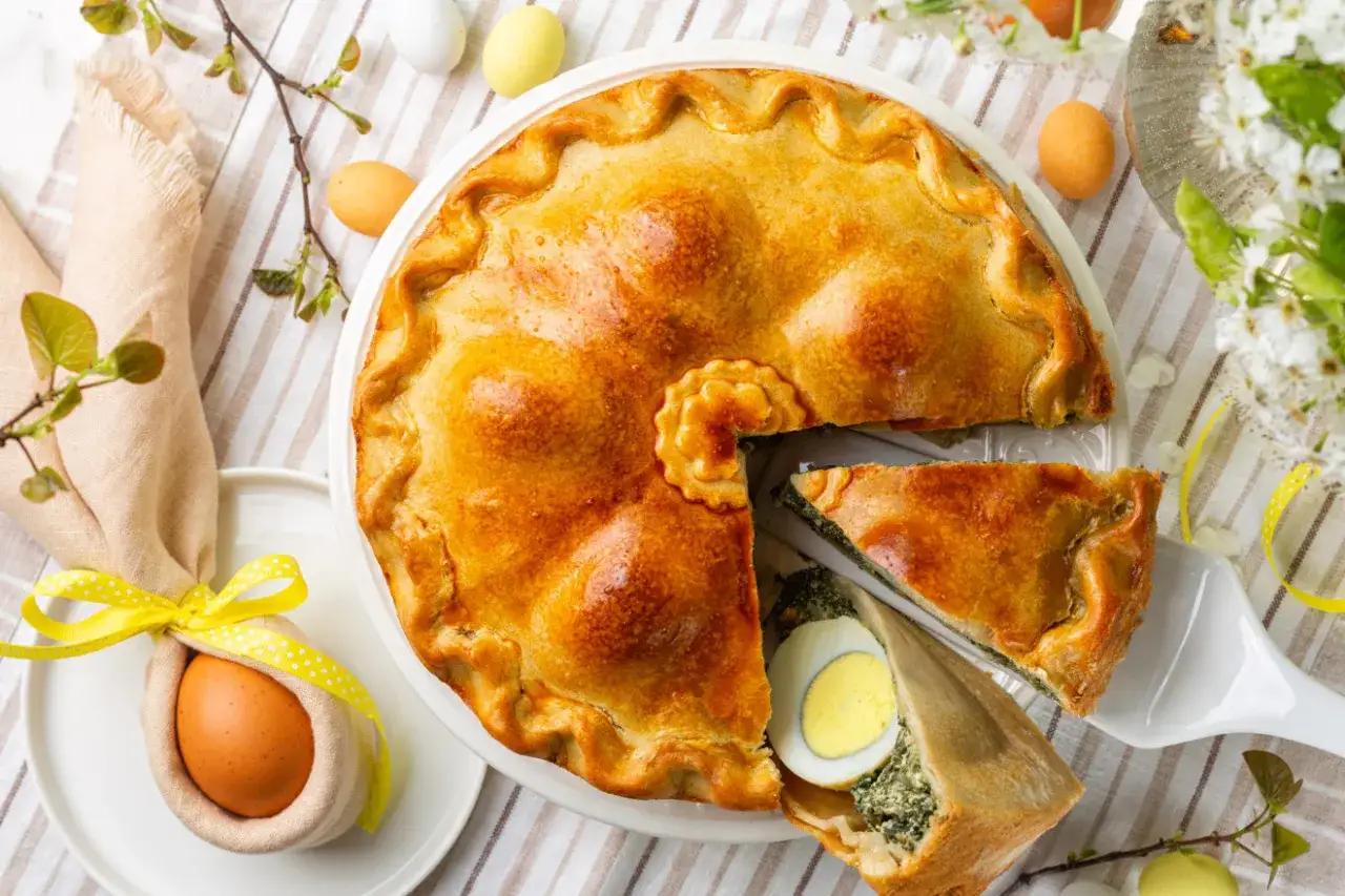 Złocista torta pasqualina z zielonym nadzieniem i jajkiem, udekorowana wielkanocnymi pisankami i gałązkami.