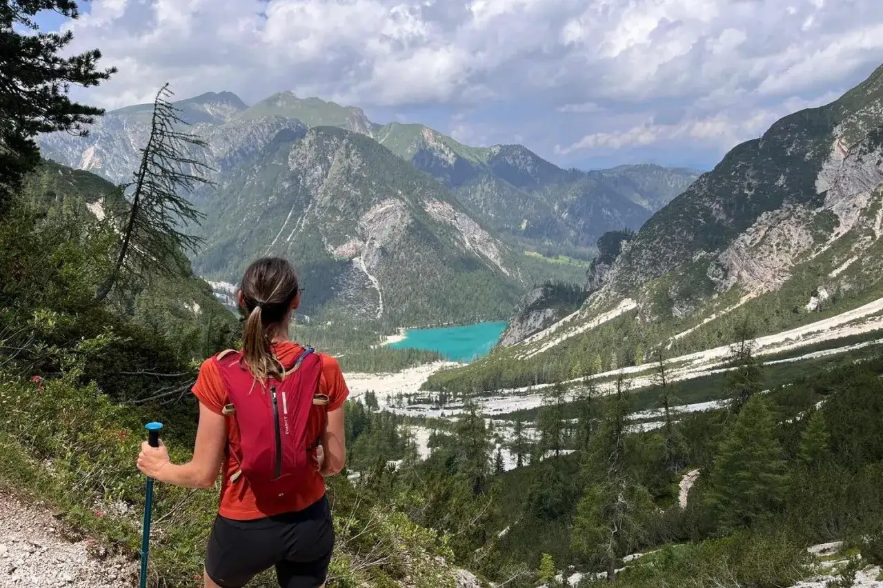 Vue panoramique sur un lac turquoise entouré de montagnes verdoyantes. Une femme en tenue de sport, sac à dos rouge et bâtons de marche, profite du paysage lors d'un entrainement trail 20 km.