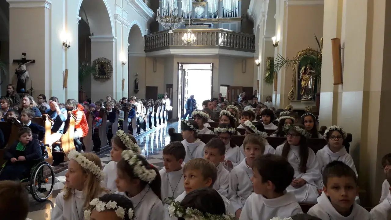 Dzieci w bieli, z wiankami na głowach, siedzą w kościele. Jeden chłopiec na wózku zastanawia się: