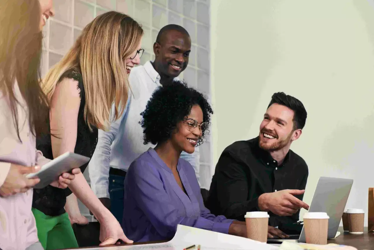 A diverse team collaborates, laughing as they review a laptop, perhaps discussing performance review challenges and examples.