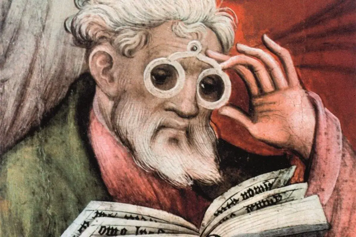 Starzec z brodą i siwymi włosami czyta księgę, używając prymitywnych okularów. To obraz z czasów, kiedy wynaleziono okulary.