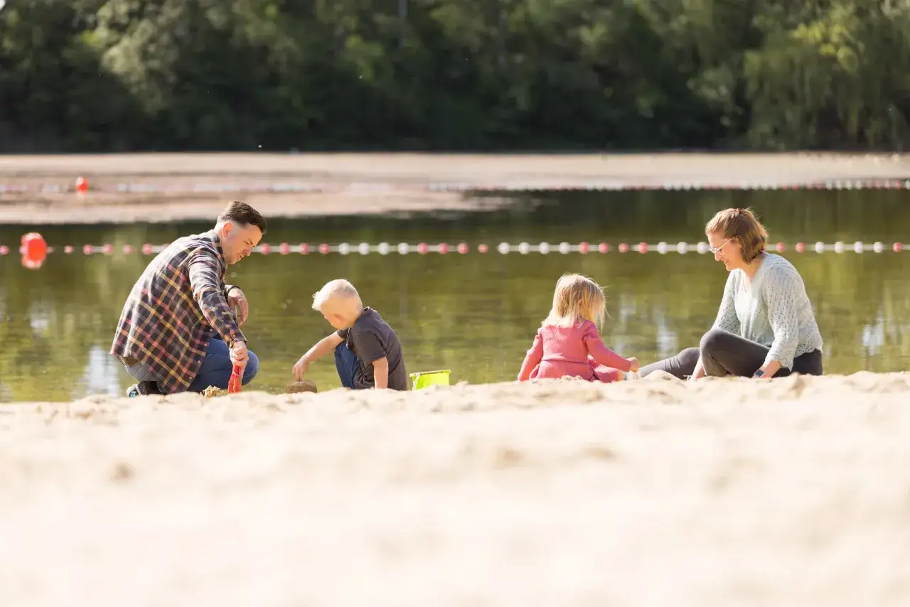 Familie spielt am Sandstrand des Badesee Mindenerwald. Vater hilft Sohn beim Sandburgen bauen, während Mutter und Tochter daneben sitzen.