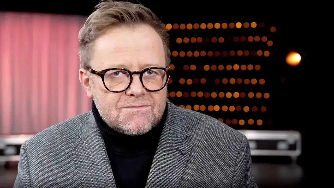Olaf Lubaszenko w okularach i marynarce, w tle światła.