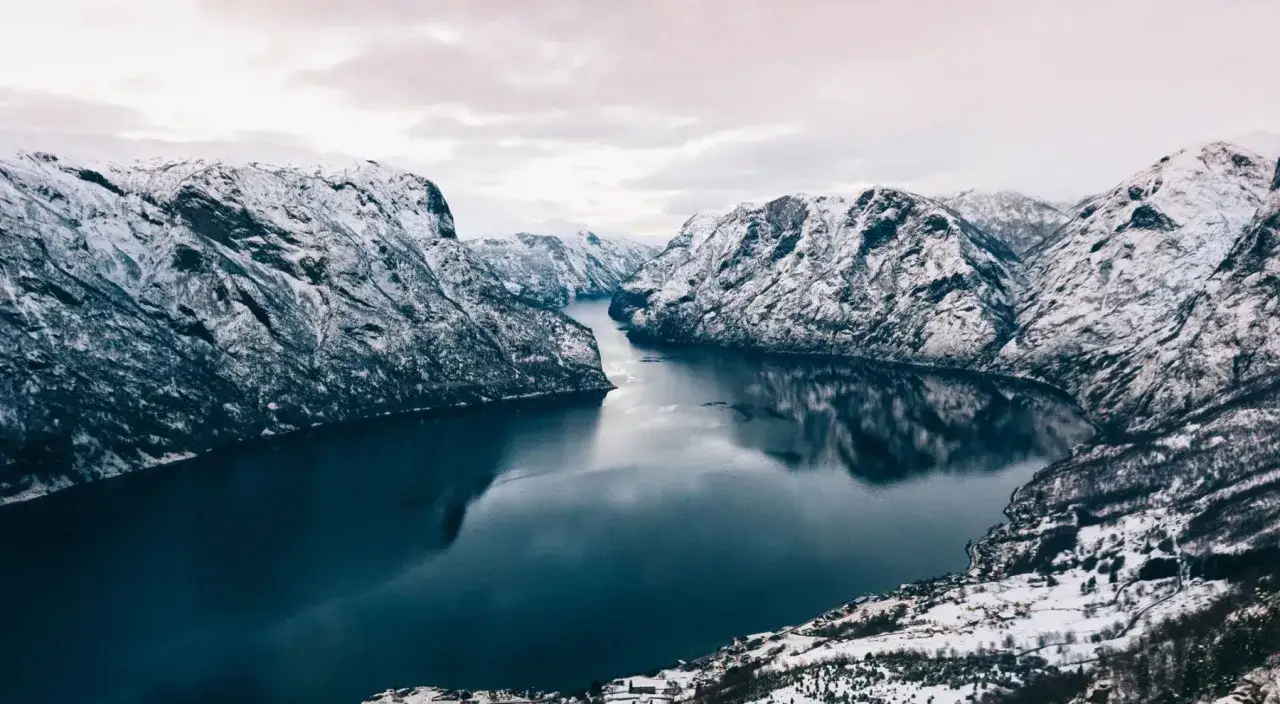 Zimowa Norwegia. Majestatyczne fiordy otoczone ośnieżonymi górami, odbijające się w spokojnej, ciemnej wodzie.
