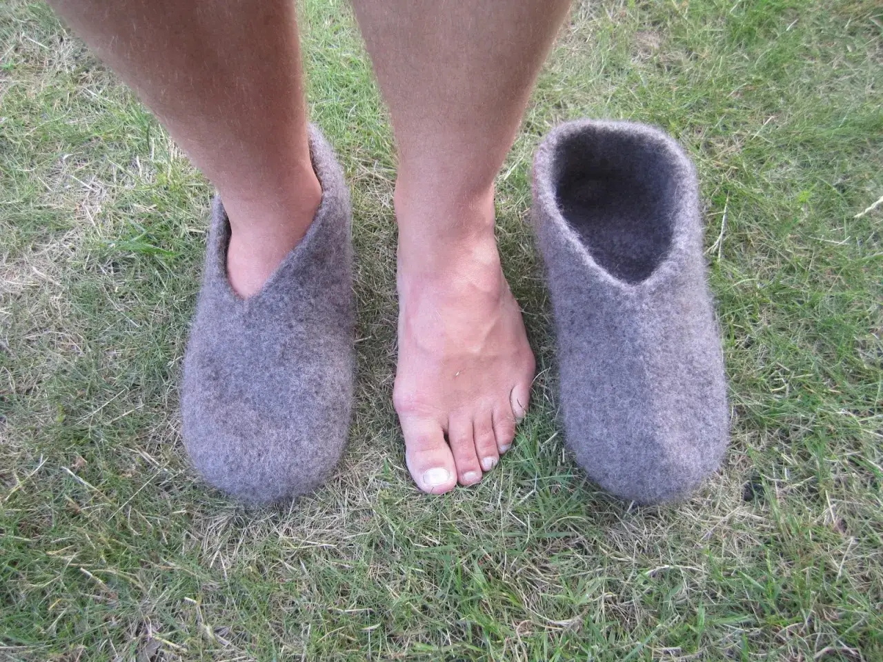 Gr&aring;, sticka tovade tofflor st&aring;r p&aring; gr&auml;set bredvid ett par bara f&ouml;tter.