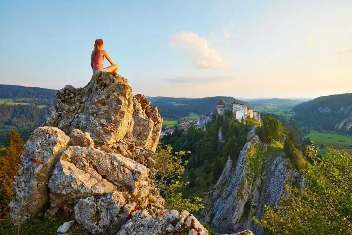 Vue panoramique d'une femme méditant sur un rocher, contemplant un château perché sur une falaise. Un paysage de la grande traversée du Jura 7 jours.