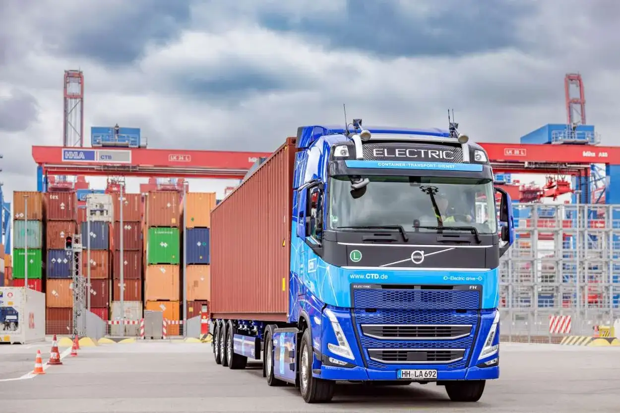 Ein elektrischer LKW mit Container beladen, im Hintergrund Kr&auml;ne und Containerstapel. Dies ist ein typischer Anblick in einem Container-Terminal.