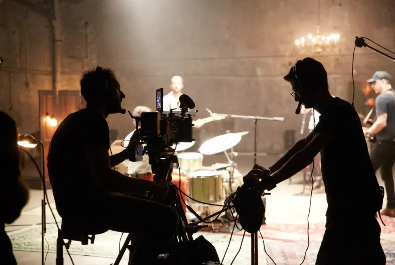 Ekipa filmowa pracuje nad teledyskiem. W tle zespół muzyczny gra na instrumentach, a operatorzy kamery rejestrują ujęcia.
