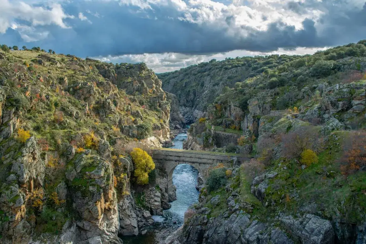 Un puente de piedra cruza un río encajonado entre las **montañas de Madrid**. El paisaje otoñal se tiñe de ocres y verdes bajo un cielo nublado.