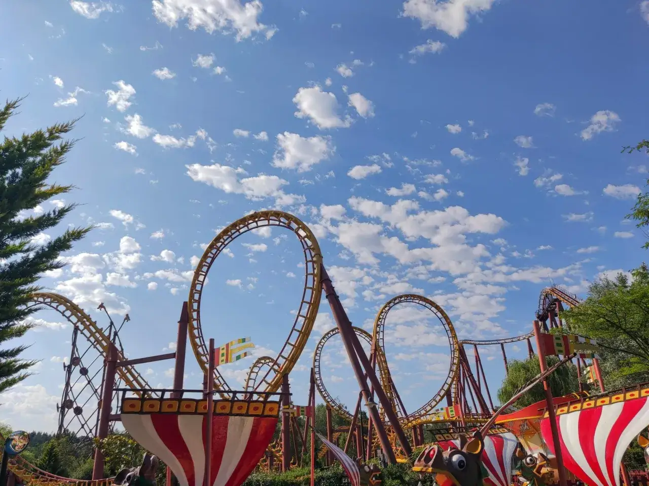 Montagnes russes jaunes et rouges dans un parc attraction, sous un ciel bleu parsemé de nuages blancs.