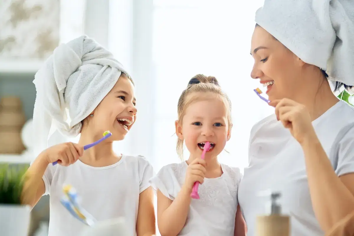 Mama i dwie córki myją zęby. Młodsza córka, która nie chce myć zębów, śmieje się, trzymając różową szczoteczkę.