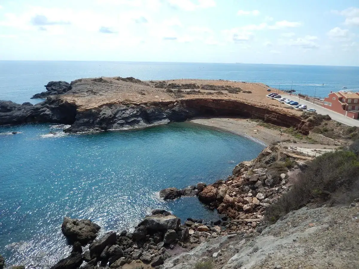 Carretera subida al faro con vistas a una cala de aguas turquesas y formaciones rocosas.