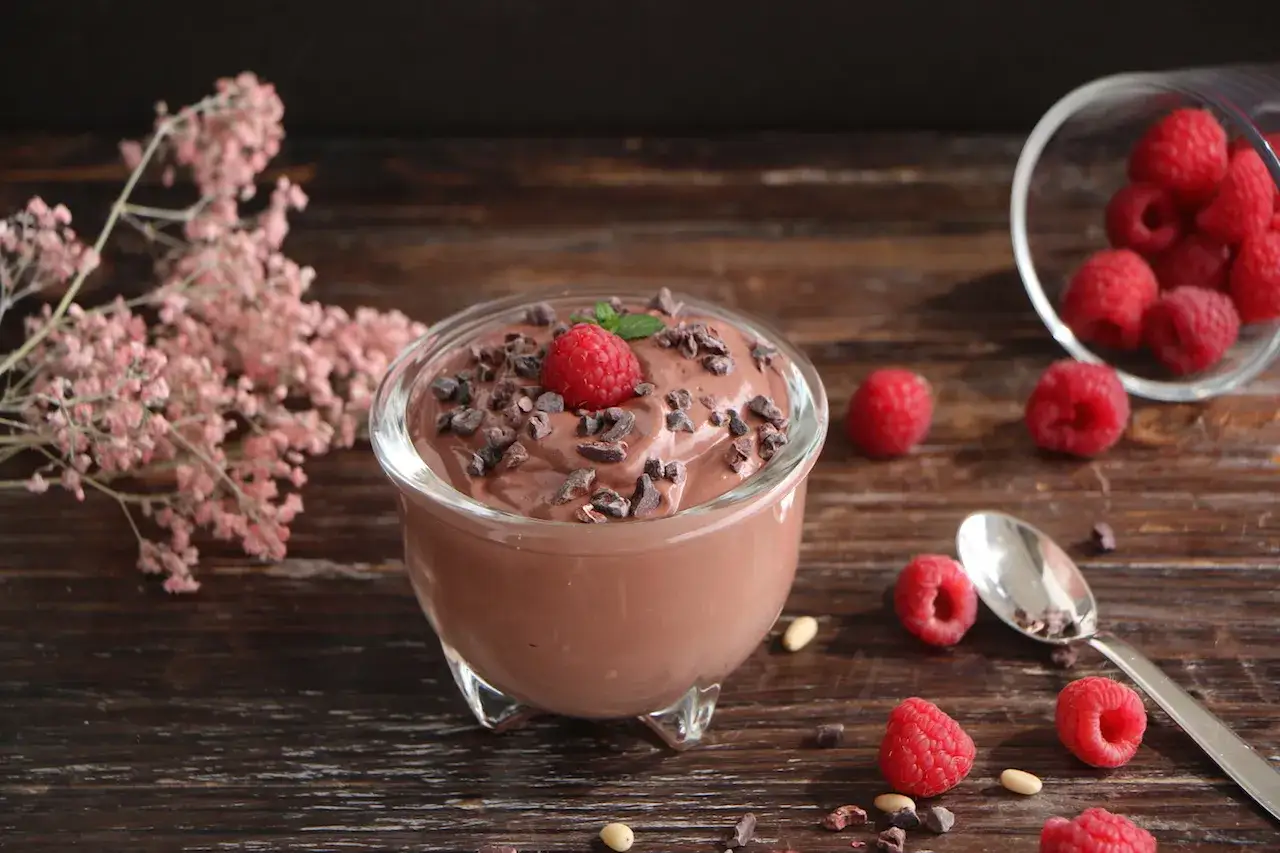 Omas schokoladenpudding mit sahne, garniert mit Himbeeren und Kakaonibs, auf einem Holztisch mit frischen Himbeeren und einer kleinen rosa Blume.