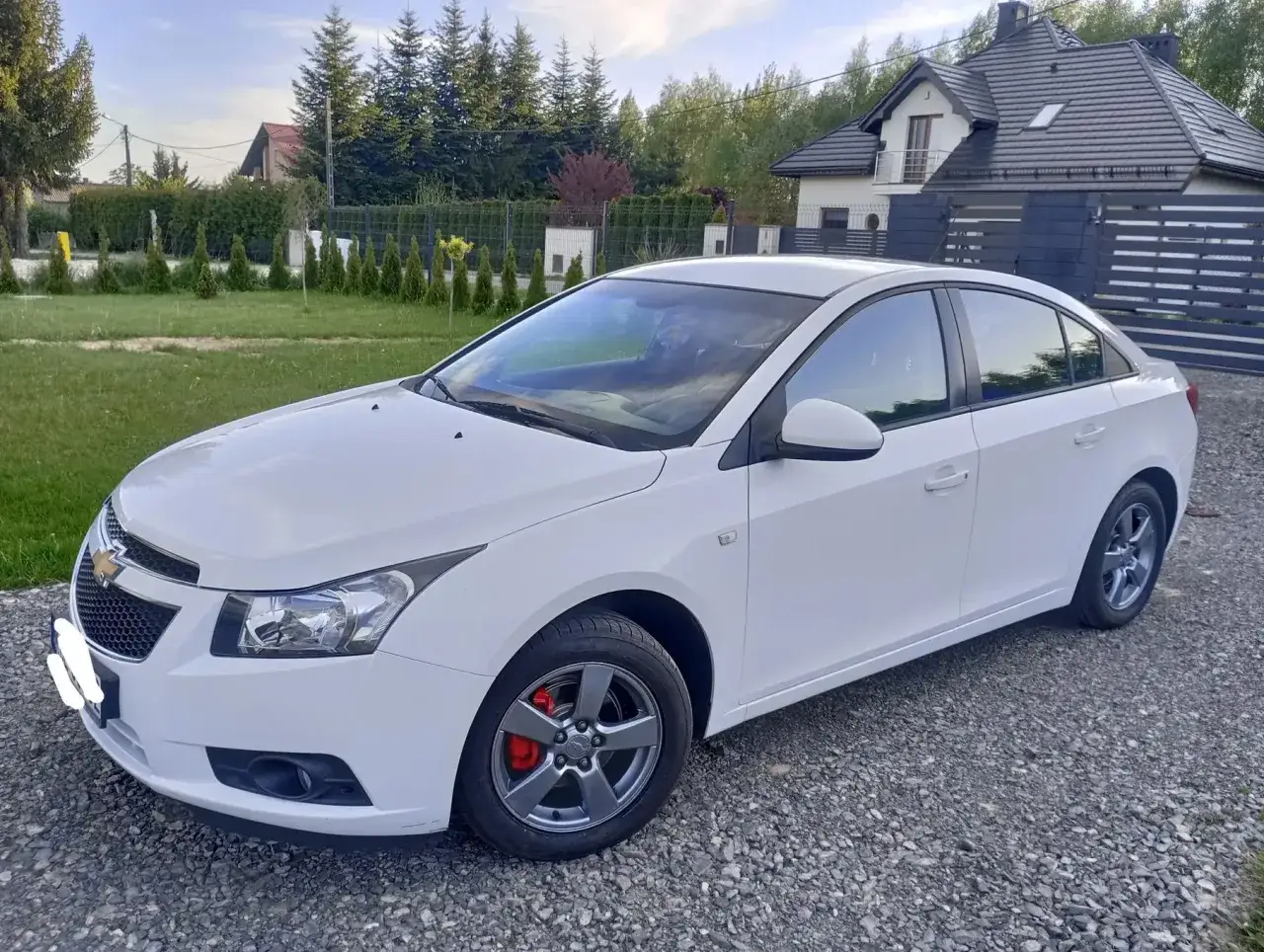 Biały Chevrolet Cruze 1.8 benzyna zaparkowany na żwirowej posesji, z zielonym trawnikiem i domem w tle.