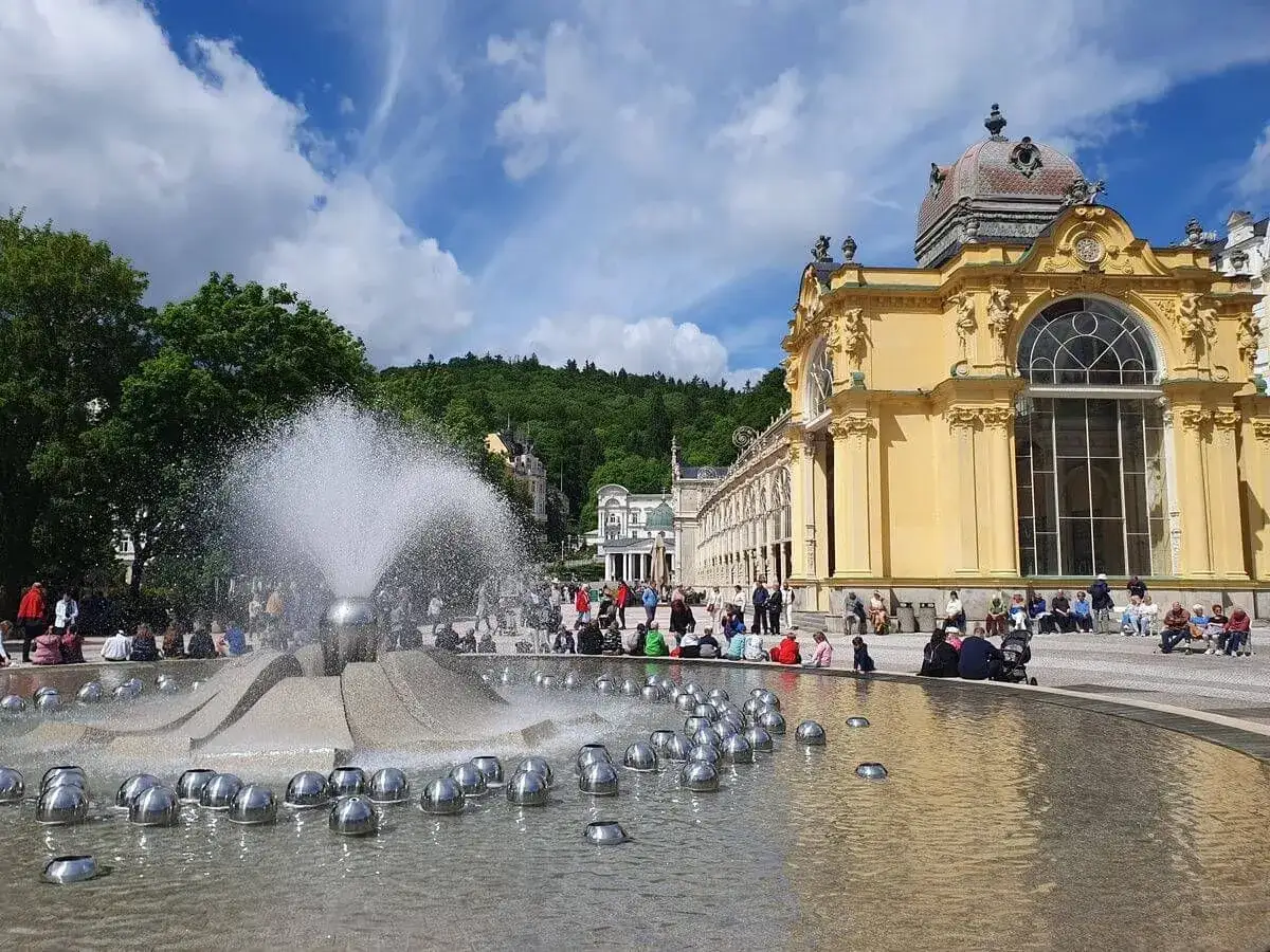Brunnen spr&uuml;ht in Mari&aacute;nsk&eacute; L&aacute;zně vor pr&auml;chtigem gelbem Geb&auml;ude. Menschen genie&szlig;en die Sonne.