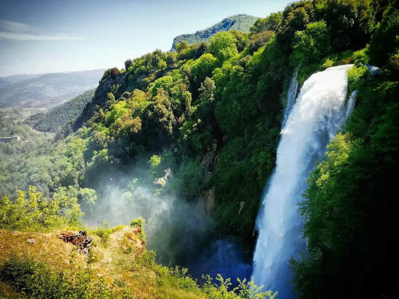 Cascata imponente nella montagna umbra, circondata da una vegetazione lussureggiante e nebbia.