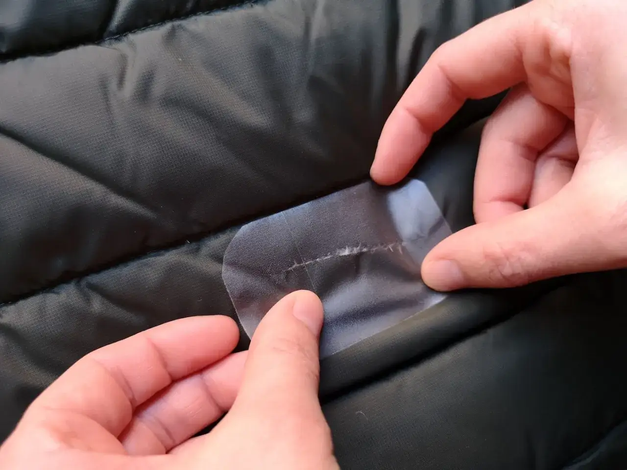 Hände reparieren eine Daunenjacke mit einem transparenten Flicken, der über einen Riss geklebt wird.