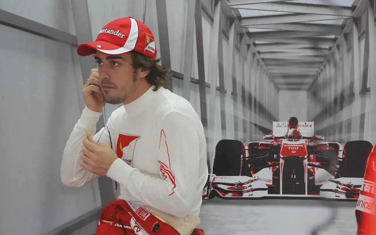 Fernando Alonso, con gorra y mono de Ferrari, se prepara para la carrera. El fondo muestra un coche de F1 rojo y blanco.