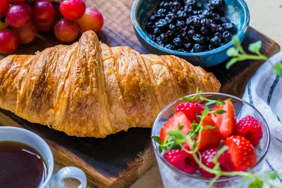 En gyllene croissant ligger på en träskärbräda, redo att baka. Bredvid finns färska bär, vindruvor och en kopp kaffe.