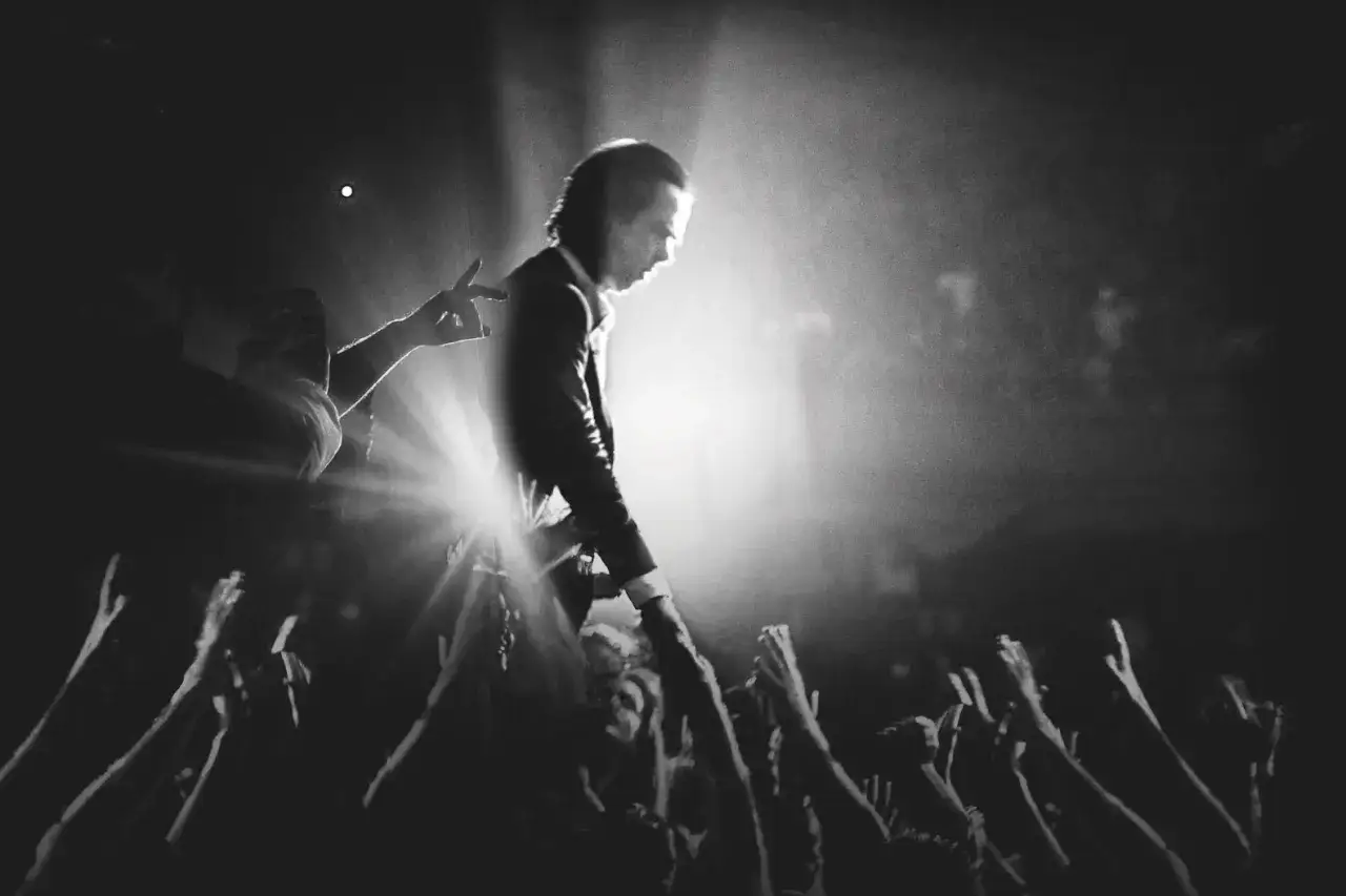 Nick Cave pochyla się nad tłumem, sięgając ręką do fanów. W świetle reflektorów widać jego charakterystyczną sylwetkę, a w powietrzu czuć energię jego utworów.