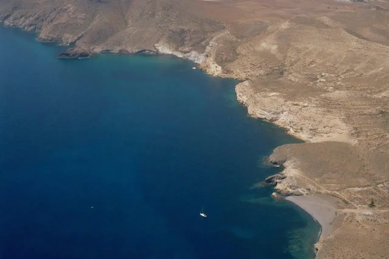 Vista aérea de la cala del Carnaje, con aguas azules y acantilados áridos. Un velero navega solitario.