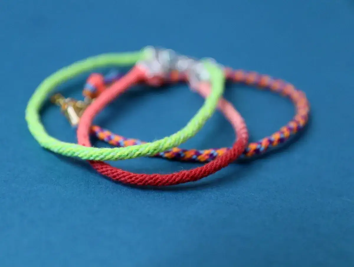 Tre färgglada kumihimo armband i neonrosa, limegrönt och rött med ett mönstrat band i bakgrunden.
