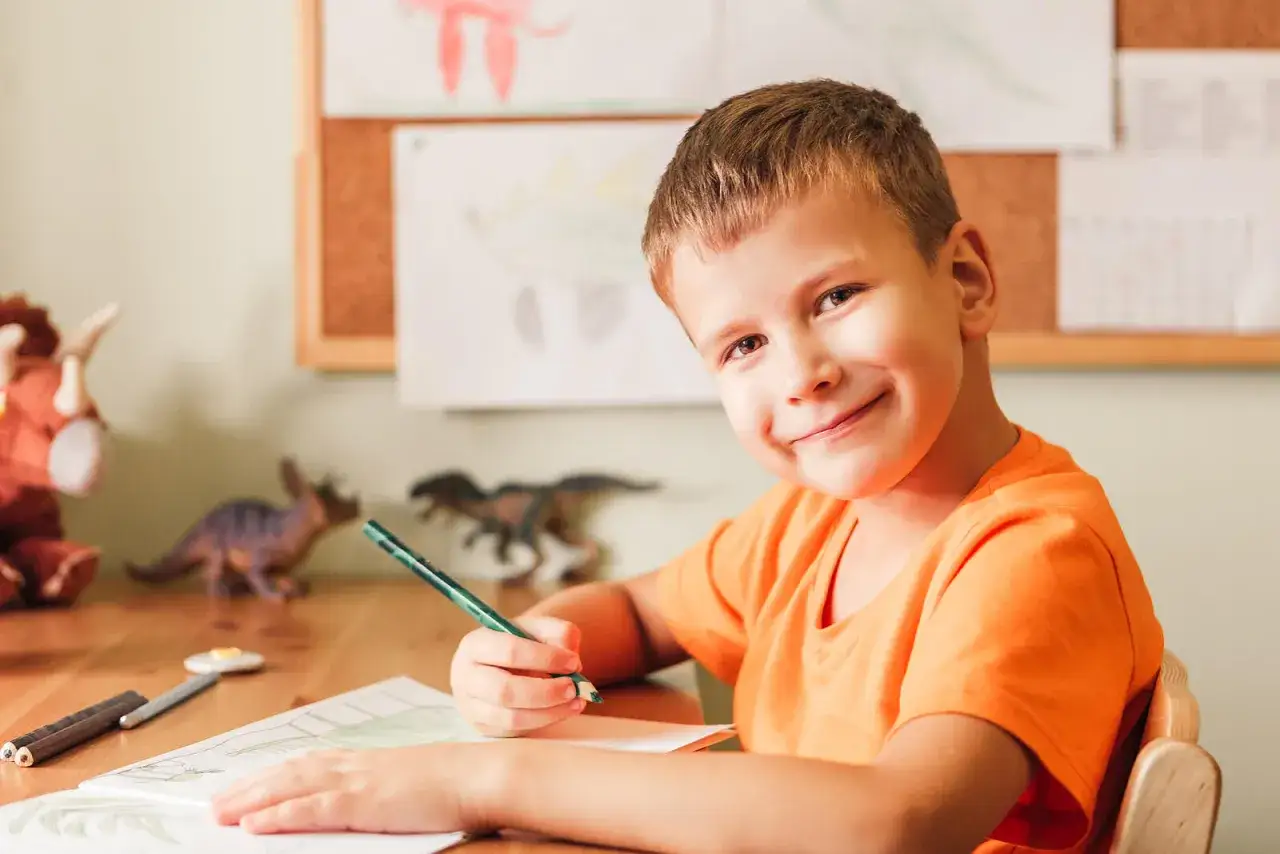 Niño sonriente practica escritura manual con un lápiz verde, rodeado de dinosaurios de juguete y dibujos.