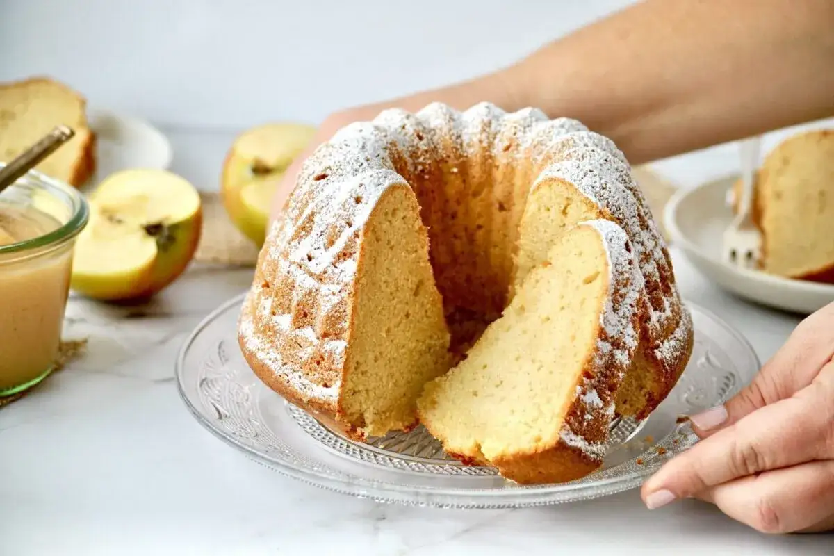 Ein **schneller saftiger Kuchen mit Apfelmus** wird auf einem Glasst&auml;nder pr&auml;sentiert, best&auml;ubt mit Puderzucker. Daneben liegen &Auml;pfel und ein Glas Apfelmus.